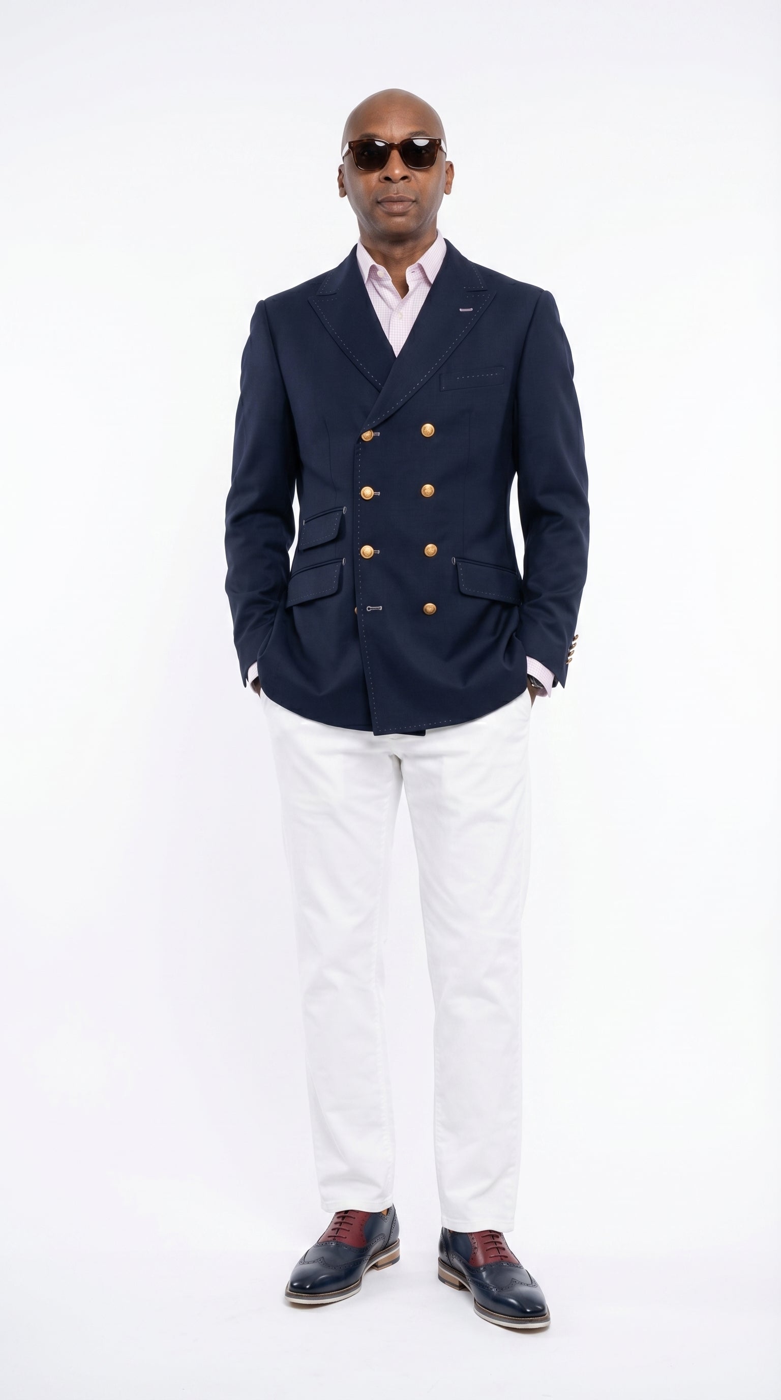 Man wearing a navy blazer with gold buttons, white shirt, and white pants on a white background