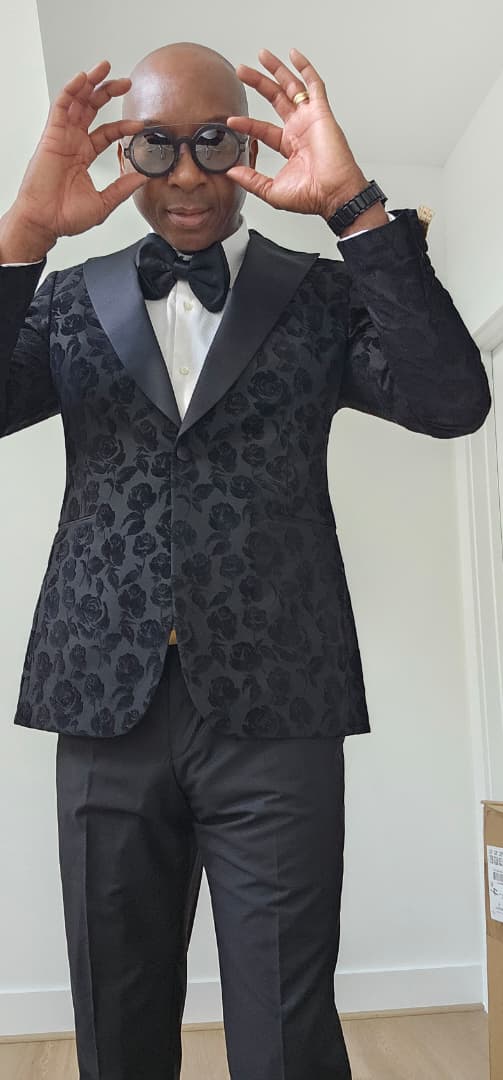Man wearing a black floral tuxedo with a white shirt and black bow tie, posing indoors.