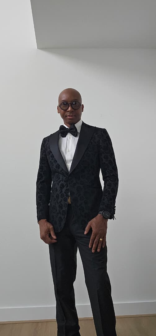Man wearing a black patterned tuxedo with a white shirt and bow tie against a plain background.