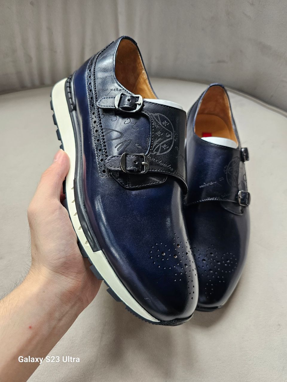 Pair of navy blue leather shoes with buckles held by a hand on a light gray surface.