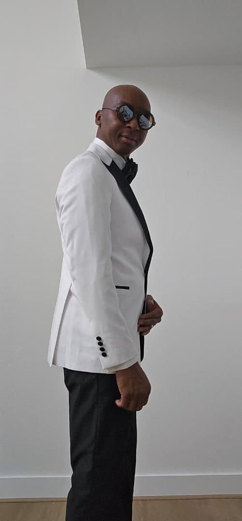 Man wearing a white blazer and black pants against a plain wall.