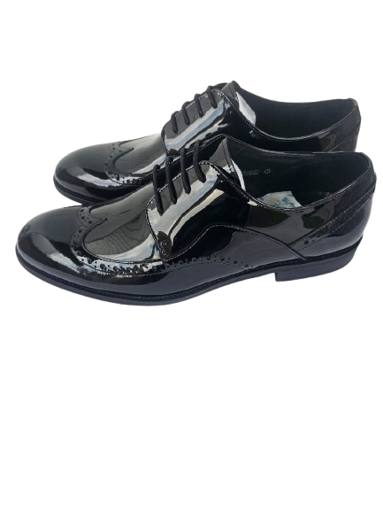 A pair of glossy black patent leather lace-up wingtip shoes with decorative brogue detailing, displayed on a white surface against a light textured wall.